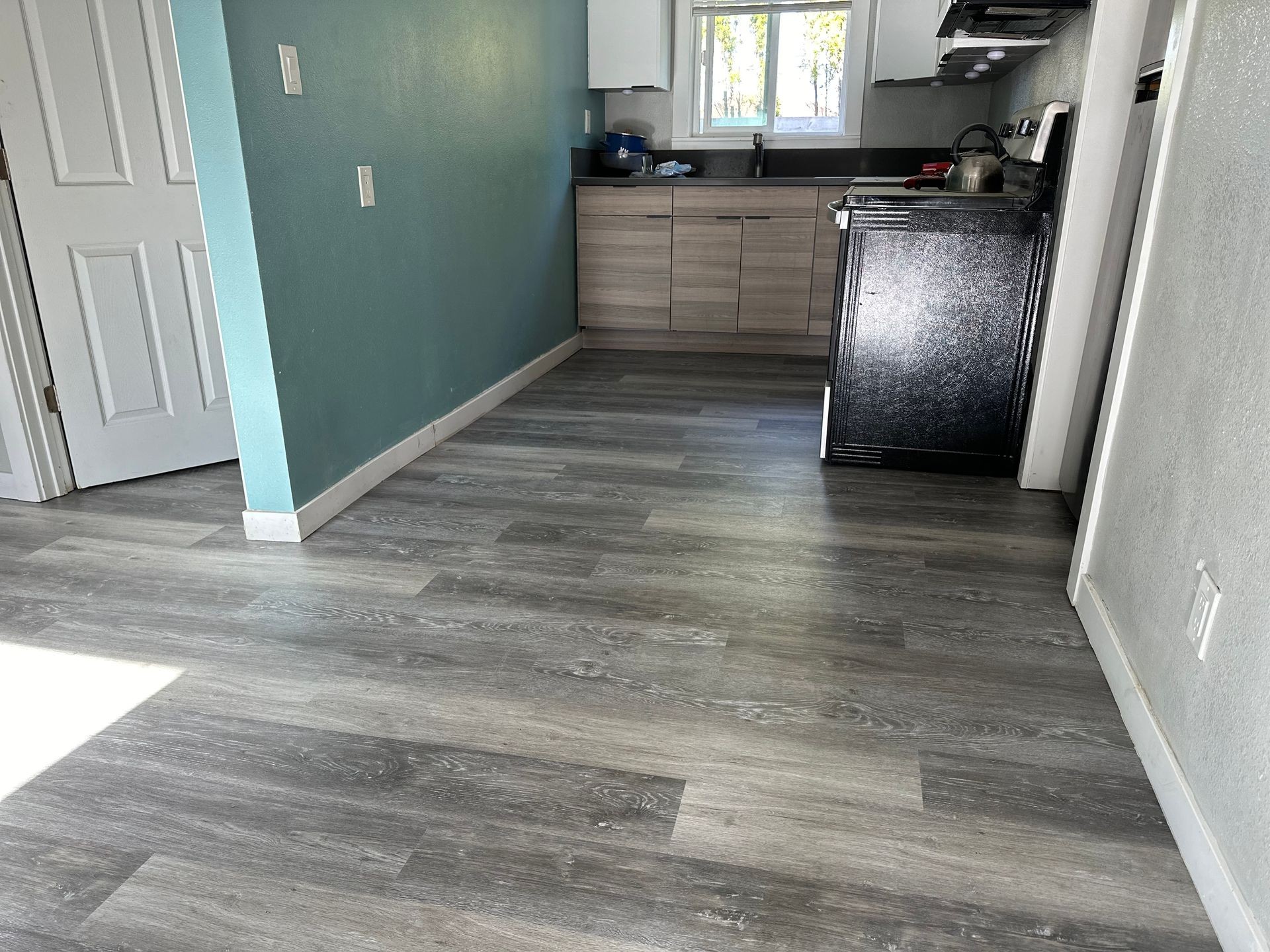 Empty kitchen with teal wall, gray wood flooring, and light wooden cabinets in sunlight.